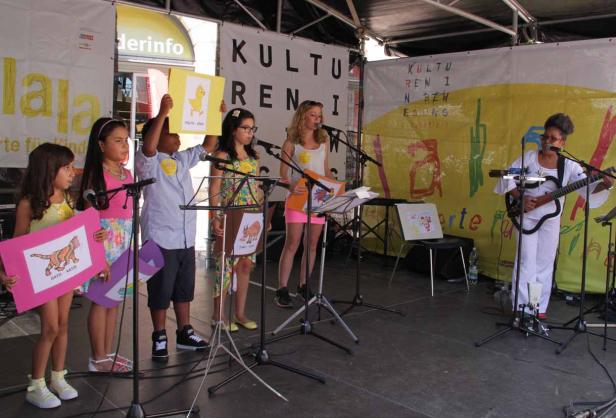 Kinder präsentieren Zeichnungen und singen auf einer Bühne, begleitet von einer Gitarristin.