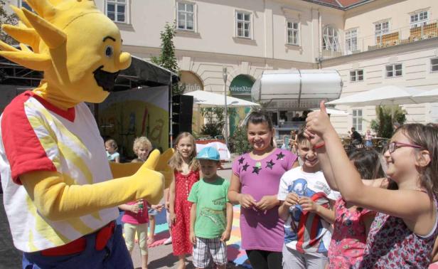 Eine Gruppe Kinder steht mit einem gelben Maskottchen vor einem Gebäude.