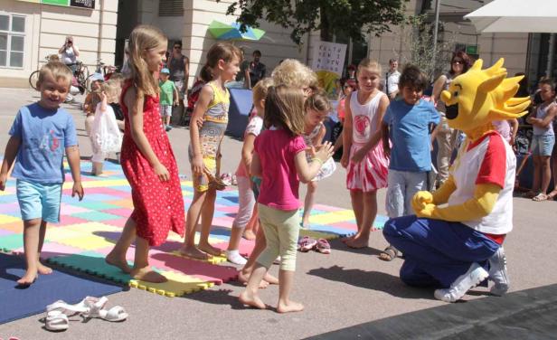 Eine Gruppe Kinder spielt barfuß mit einem Maskottchen auf einer bunten Matte.