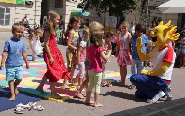 Eine Gruppe Kinder spielt mit einem Sonnenmaskottchen auf einer bunten Matte.