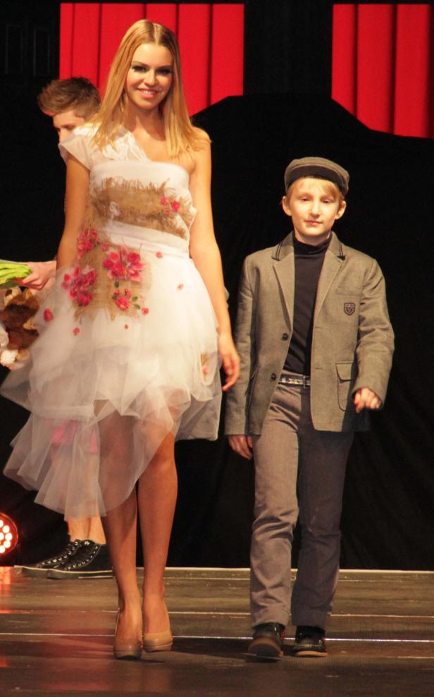 Eine junge Frau in einem weißen Kleid und ein Junge mit Schirmmütze laufen auf einem Laufsteg.