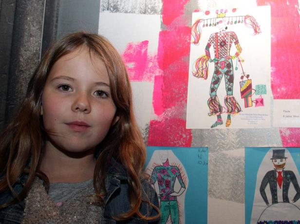 Ein junges Mädchen steht vor einer Wand mit Kinderzeichnungen von Kostümen.