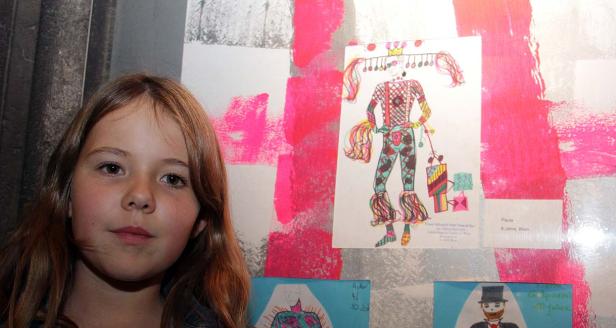 Ein Mädchen steht vor einer Wand mit Kinderzeichnungen, darunter ein Entwurf für ein Karnevalskostüm.