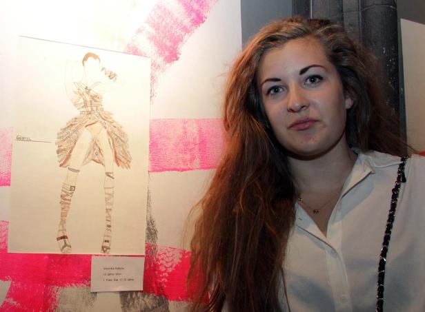 Eine junge Frau steht neben einer Zeichnung eines Gürtel-Kleides von Veronika Kobma.
