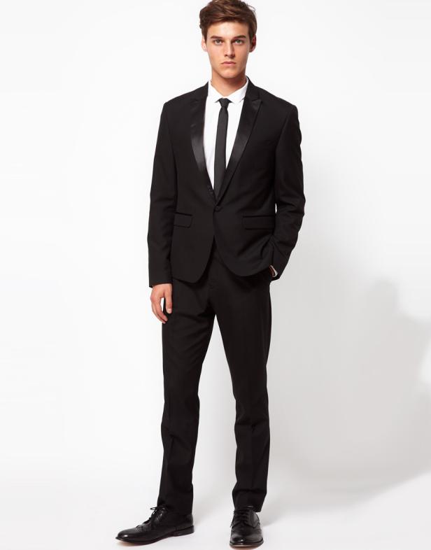 Ein Mann in einem schwarzen Anzug mit Krawatte posiert vor einem weißen Hintergrund.