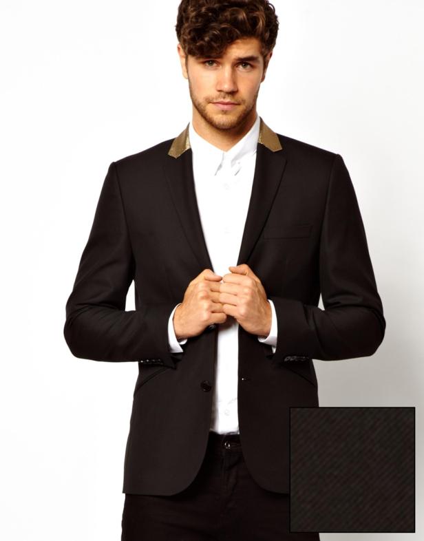 Ein Mann mit lockigem Haar trägt ein schwarzes Sakko mit goldenem Kragen über einem weißen Hemd.