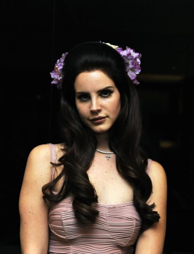 Lana Del Rey mit Blumen im Haar und einem rosa Kleid vor einem dunklen Hintergrund.