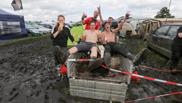 Eine Gruppe von Festivalbesuchern sitzt in einem Anhänger inmitten von Schlamm und Zelten.