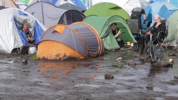 Zwei Männer und eine Frau sitzen vor ihren Zelten auf einem schlammigen Festivalgelände.