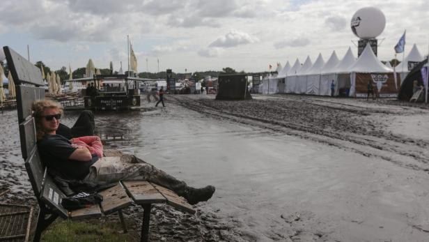 Ein Mann mit Sonnenbrille entspannt auf einer Bank inmitten des schlammigen Geländes des Wacken Open Airs.
