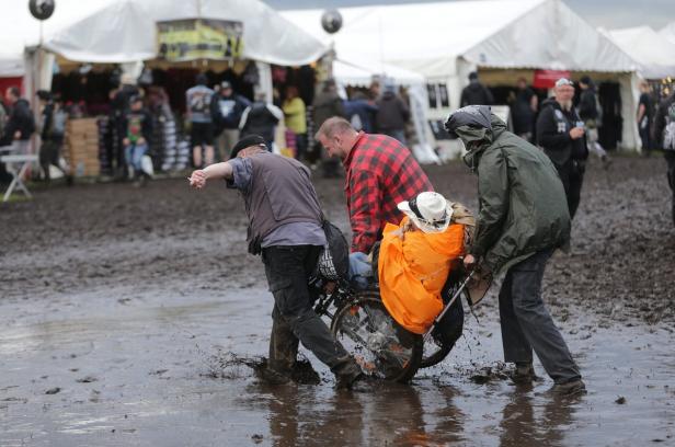 Zwei Männer schieben eine Person im Rollstuhl durch den schlammigen Boden eines Festivals.