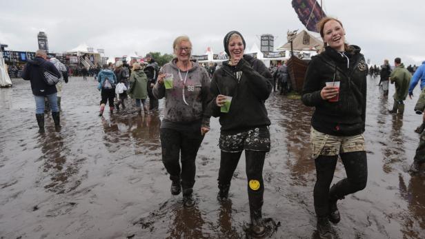 Drei Frauen waten mit Getränken durch den Schlamm auf einem Festivalgelände.
