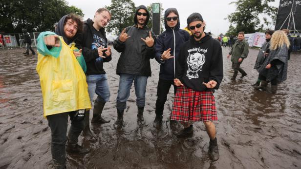 Eine Gruppe junger Männer posiert in Gummistiefeln und Regenkleidung auf einem schlammigen Festivalgelände.