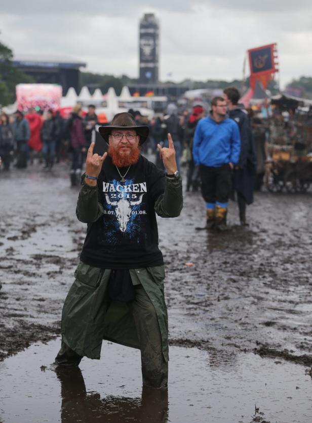 Ein Mann mit Hut und Wacken-T-Shirt steht im Schlamm und zeigt die Pommesgabel.