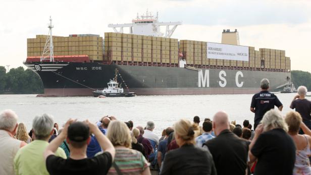 Die „MSC Zoe“ fährt auf der Elbe, begleitet von einem Schlepper, während Zuschauer am Ufer stehen.