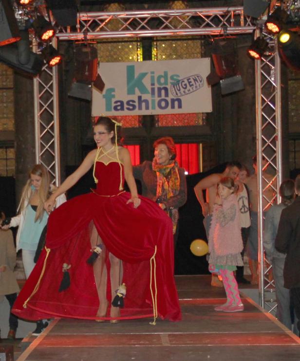 Eine Frau in einem roten Kleid mit Puppen auf einer Bühne bei einer „Kids in Fashion“-Veranstaltung.