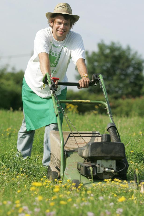 Ein Mann mit Hut schiebt einen Rasenmäher über eine blühende Wiese.