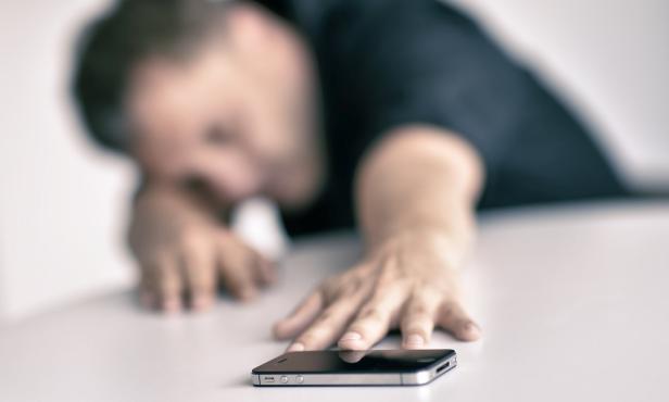 Ein Mann liegt erschöpft auf einem Tisch, sein Handy in Reichweite.