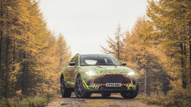 Ein Aston Martin DBX mit auffälliger Tarnlackierung steht auf einer unbefestigten Straße in einem Wald.