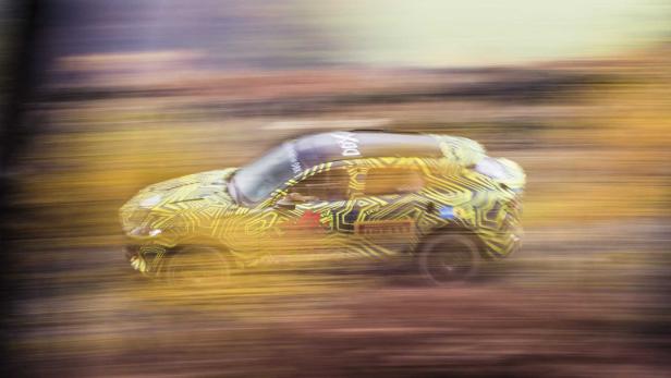Ein gelb gemusterter Aston Martin DBX fährt schnell über eine unbefestigte Straße.