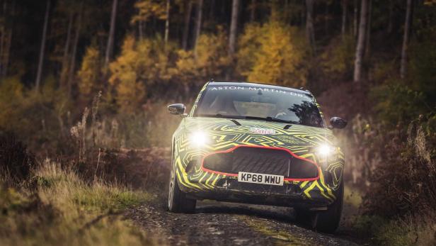 Ein getarnter Aston Martin DBX fährt auf einer Schotterstraße durch einen Wald.