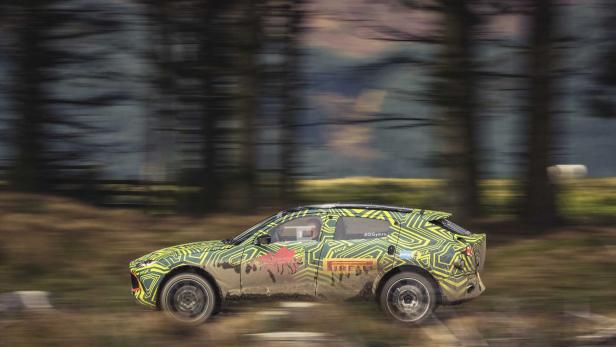 Ein getarnter Aston Martin DBX fährt schnell auf einer unbefestigten Straße.