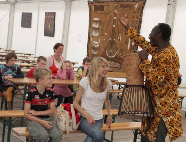 Ein Mann mit einem traditionellen Hemd spielt eine Trommel vor einer Gruppe von Kindern.