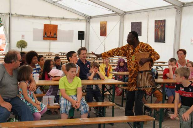 Ein Mann in traditioneller Kleidung präsentiert eine Trommel vor einer Gruppe von Kindern.