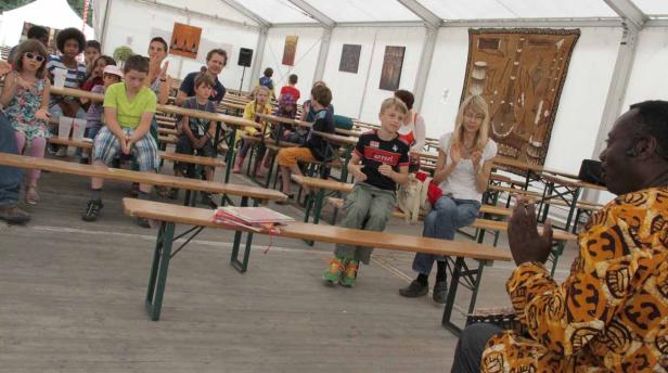 Eine Gruppe von Kindern und Erwachsenen sitzt auf Bänken und hört einem Mann in einem gemusterten Hemd zu.