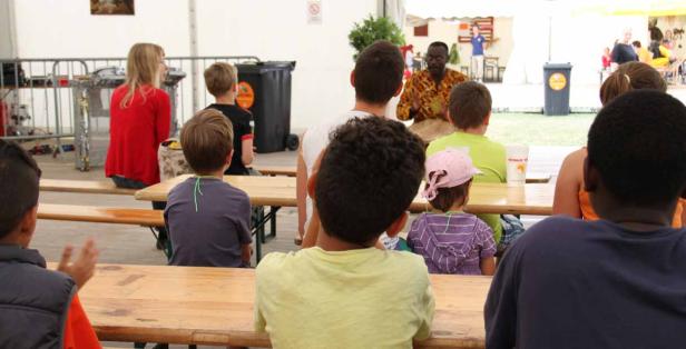 Eine Gruppe von Kindern sitzt auf Bänken und schaut einem Mann beim Trommeln zu.