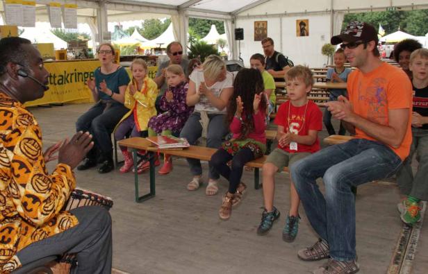 Ein Mann mit einem Mikrofon spielt Trommel vor einer Gruppe von Kindern und Erwachsenen.