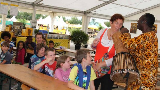 Eine Gruppe Kinder sitzt an einem Tisch, während ein Mann ihnen eine Trommel vorführt.