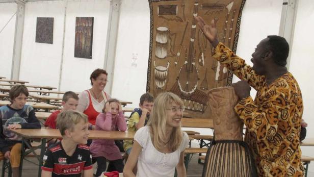 Ein Mann in gemustertem Hemd spricht vor einer Gruppe von Kindern und Erwachsenen über afrikanische Musikinstrumente.