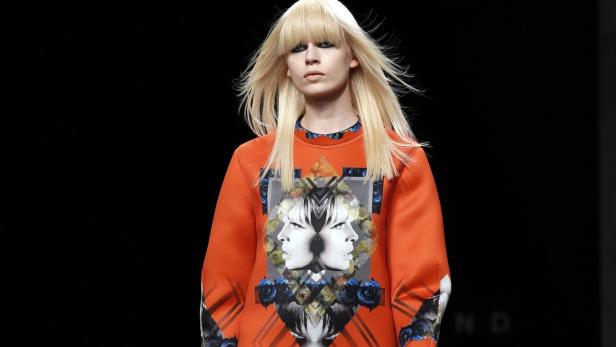 Eine blonde Frau mit Pony trägt einen orangefarbenen Pullover mit einem Gesicht-Motiv.