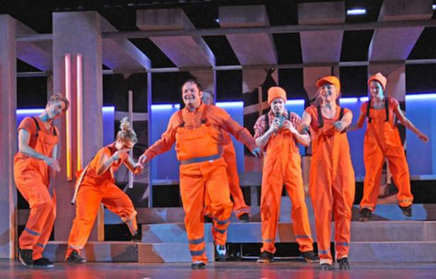 Eine Gruppe von Darstellern in orangefarbenen Overalls auf einer Bühne.