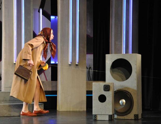 Eine Frau in Mantel und Kopftuch steht auf einer Bühne neben Lautsprechern.