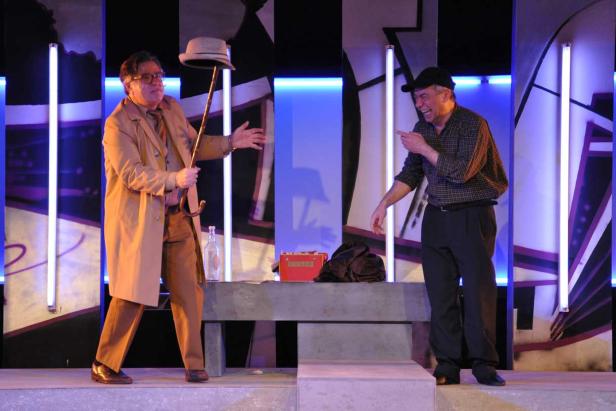 Zwei Männer stehen auf einer Bühne, einer hält einen Hut an einem Stock, der andere lacht und zeigt darauf.