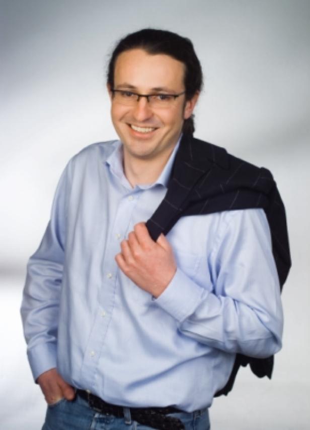 Rainer Friedl, diamond:dogs group-Board Ein lächelnder Mann mit Brille und Sakko über der Schulter steht vor einem grauen Hintergrund.