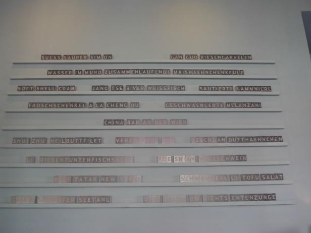 Eine Wand mit Schildern, die chinesische Gerichte auf Deutsch und Englisch auflisten.