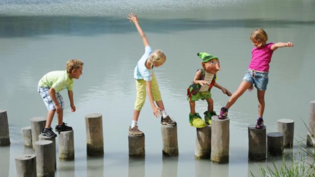 Kinder und eine Puppe balancieren auf Baumstämmen im Wasser.