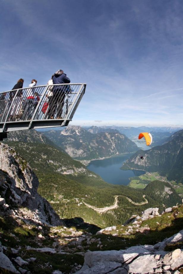 Menschen stehen auf einer Aussichtsplattform mit Blick auf einen See und einen Paraglider.