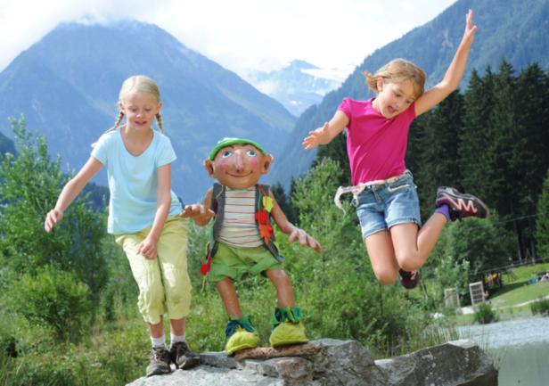 Zwei Mädchen posieren mit einer lebensgroßen Puppe vor einer Bergkulisse.