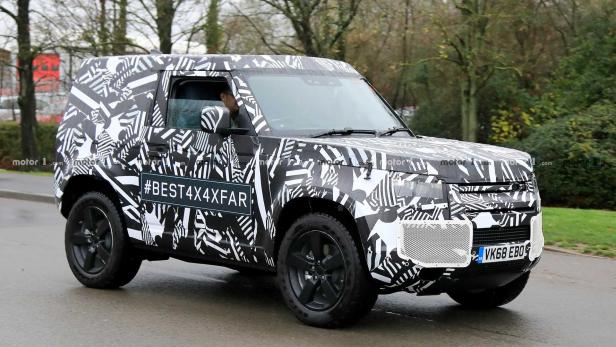Ein Prototyp des Land Rover Defender mit auffälliger Tarnung fährt auf einer Straße.