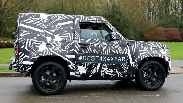 Ein mit einem schwarz-weißen Muster getarnter Land Rover Defender fährt auf einer Straße.