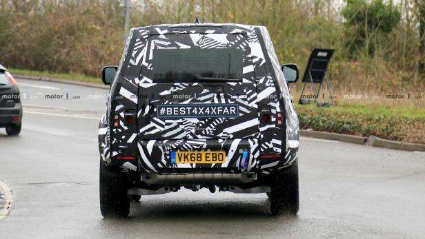 Ein Prototyp eines Land Rover Defender mit auffälliger Tarnung auf einer nassen Straße.