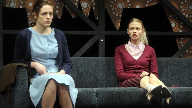 Zwei Frauen sitzen auf einer grauen Couch vor einer Bühnenkulisse.