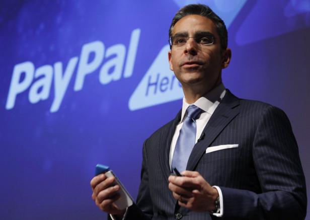 Ein Mann im Anzug hält ein Gerät vor einem PayPal-Logo.