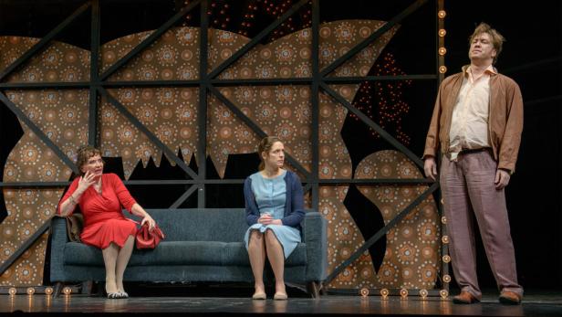 Auf einer Theaterbühne sitzen zwei Frauen auf einem Sofa, während ein Mann daneben steht.