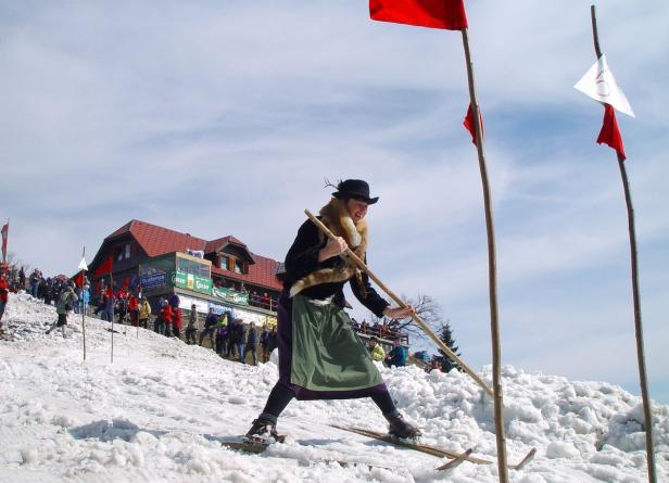 Eine Frau in Tracht fährt mit Holzskiern einen schneebedeckten Hang hinunter.