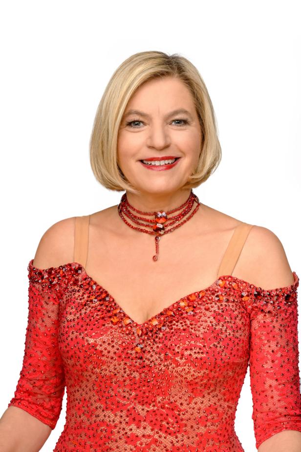 Eine lächelnde Frau mit blonden Haaren trägt ein rotes Kleid und eine rote Halskette.